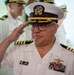 A U.S. Navy officer is rendering a salute while wearing a white service dress uniform with a combination cover. His name tag reads 'Jerome Catamotang' and he displays multiple service ribbons, a surface warfare insignia, and captain-level rank insignia. A second Navy officer in the background also appears to be saluting.