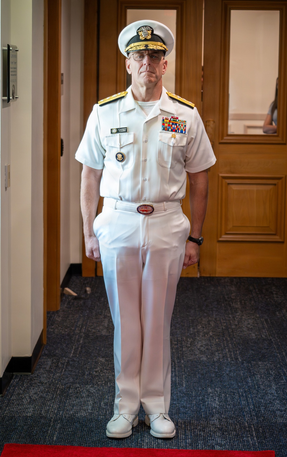 A senior U.S. Navy officer stands at attention in a formal indoor setting wearing a white service dress uniform. The uniform displays multiple rows of service ribbons, gold shoulder boards indicating flag officer rank, and a name tag. The officer is posed formally in what appears to be an official or ceremonial context.