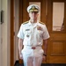 A senior U.S. Navy officer stands at attention in a formal indoor setting wearing a white service dress uniform. The uniform displays multiple rows of service ribbons, gold shoulder boards indicating flag officer rank, and a name tag. The officer is posed formally in what appears to be an official or ceremonial context.