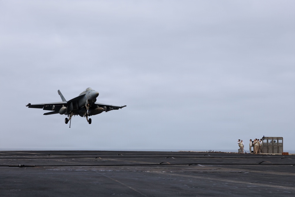 A carrier-based jet fighter, consistent with an F/A-18 Hornet or Super Hornet, is captured in a low-altitude approach or just-launched position above the flight deck of an aircraft carrier. The landing gear is fully extended, suggesting an arrested landing approach. A group of personnel in tan/khaki uniforms are visible on the right side of the deck near what appears to be a deck-edge structure.
