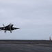 A carrier-based jet fighter, consistent with an F/A-18 Hornet or Super Hornet, is captured in a low-altitude approach or just-launched position above the flight deck of an aircraft carrier. The landing gear is fully extended, suggesting an arrested landing approach. A group of personnel in tan/khaki uniforms are visible on the right side of the deck near what appears to be a deck-edge structure.