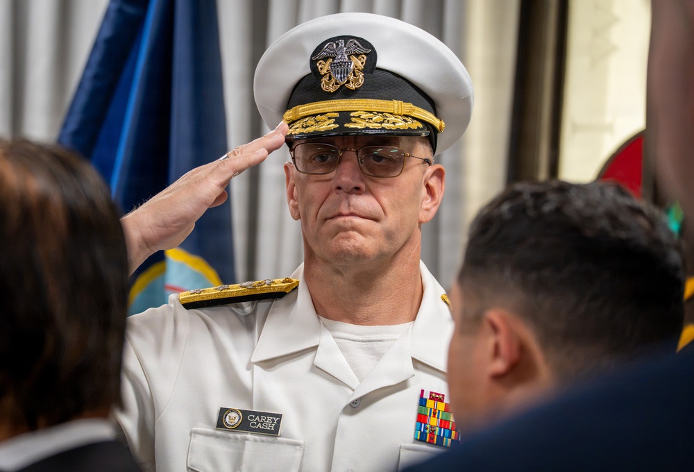A U.S. Navy officer identified by his name tag as 'Carey Cash' is rendering a salute during what appears to be a formal military ceremony. He is wearing a white dress uniform with gold shoulder boards indicating flag officer rank, along with multiple service ribbons. The setting appears to be an indoor official function with flags visible in the background.