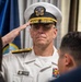 A U.S. Navy officer identified by his name tag as 'Carey Cash' is rendering a salute during what appears to be a formal military ceremony. He is wearing a white dress uniform with gold shoulder boards indicating flag officer rank, along with multiple service ribbons. The setting appears to be an indoor official function with flags visible in the background.