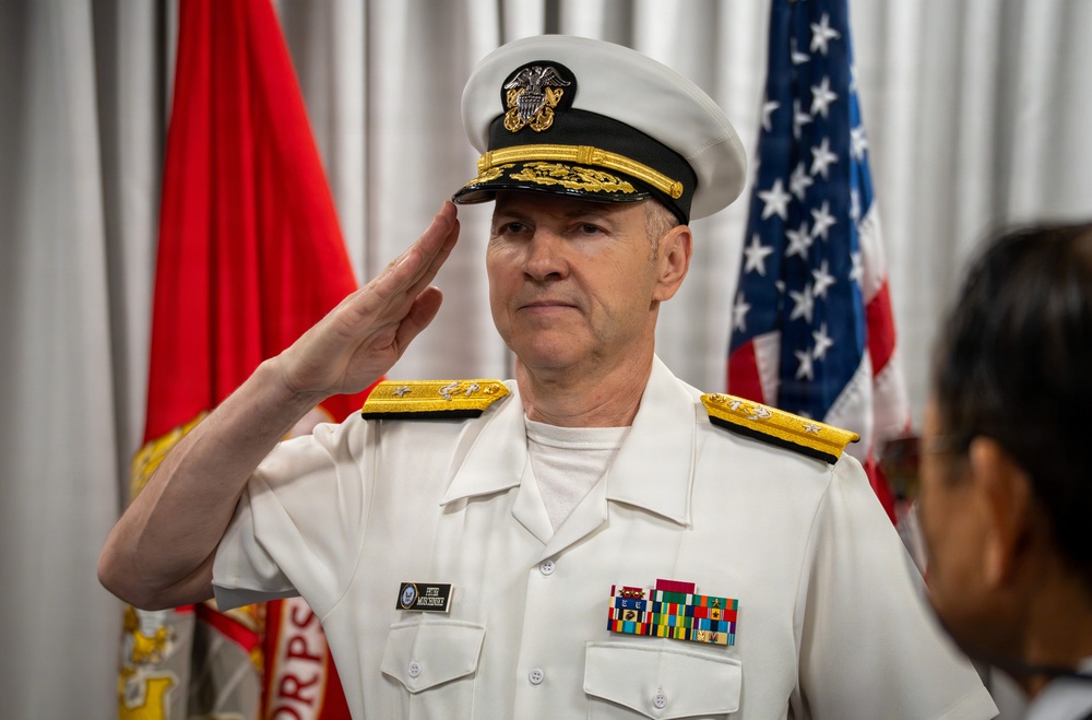 A senior U.S. Navy officer in white service dress uniform is rendering a formal salute, likely during a military ceremony. The officer displays multiple service ribbons and medals on his chest, and wears shoulder boards indicating flag officer (admiral) rank. An American flag and a Marine Corps flag are visible in the background.