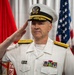 A senior U.S. Navy officer in white service dress uniform is rendering a formal salute, likely during a military ceremony. The officer displays multiple service ribbons and medals on his chest, and wears shoulder boards indicating flag officer (admiral) rank. An American flag and a Marine Corps flag are visible in the background.