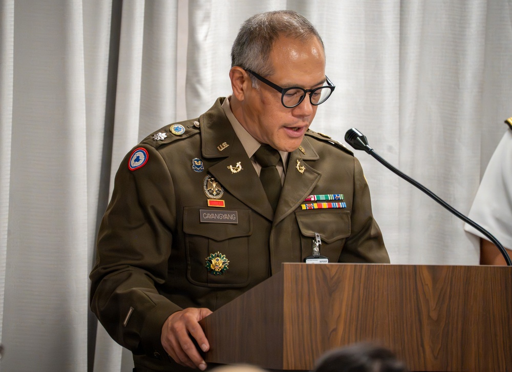 A U.S. Army officer identified by nameplate as 'CAYANGYANG' is speaking at a wooden podium with a microphone. He is wearing the Army Service Uniform (ASU) in olive green with numerous badges, ribbons, and insignia. The setting appears to be an indoor formal military ceremony or official event.