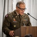 A U.S. Army officer identified by nameplate as 'CAYANGYANG' is speaking at a wooden podium with a microphone. He is wearing the Army Service Uniform (ASU) in olive green with numerous badges, ribbons, and insignia. The setting appears to be an indoor formal military ceremony or official event.