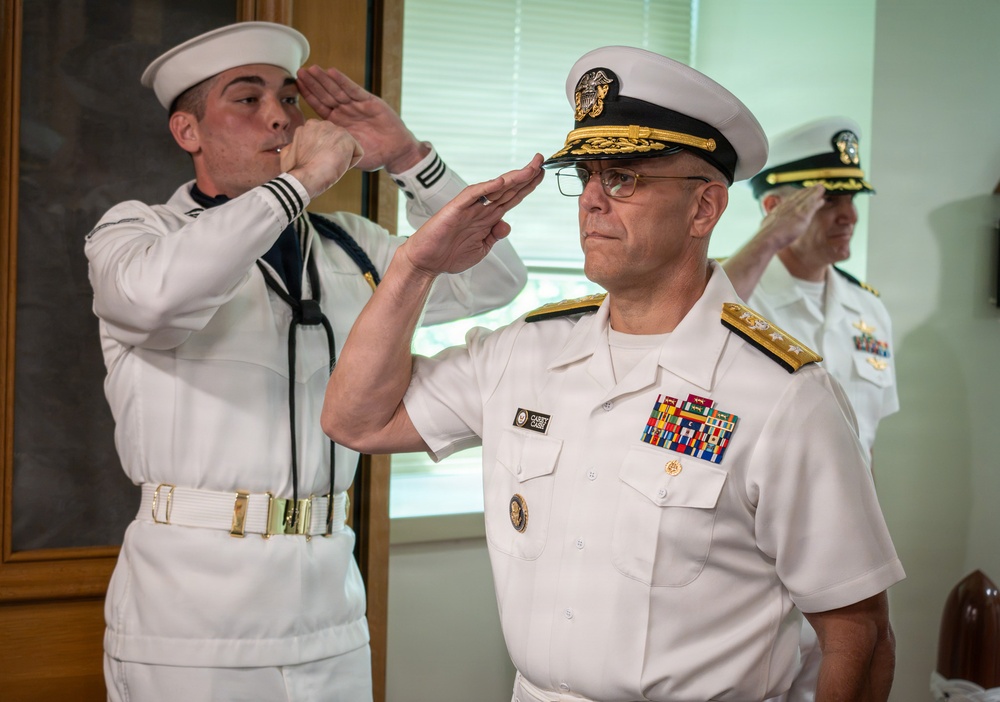 The image shows a formal U.S. Navy military ceremony with personnel in dress white uniforms rendering salutes. A senior officer in the foreground, identifiable by gold shoulder boards with stars indicating flag officer rank and an extensive row of service ribbons, leads the salute. At least three naval personnel are visible, suggesting an official ceremony such as a change of command, retirement, or formal review.