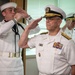 The image shows a formal U.S. Navy military ceremony with personnel in dress white uniforms rendering salutes. A senior officer in the foreground, identifiable by gold shoulder boards with stars indicating flag officer rank and an extensive row of service ribbons, leads the salute. At least three naval personnel are visible, suggesting an official ceremony such as a change of command, retirement, or formal review.