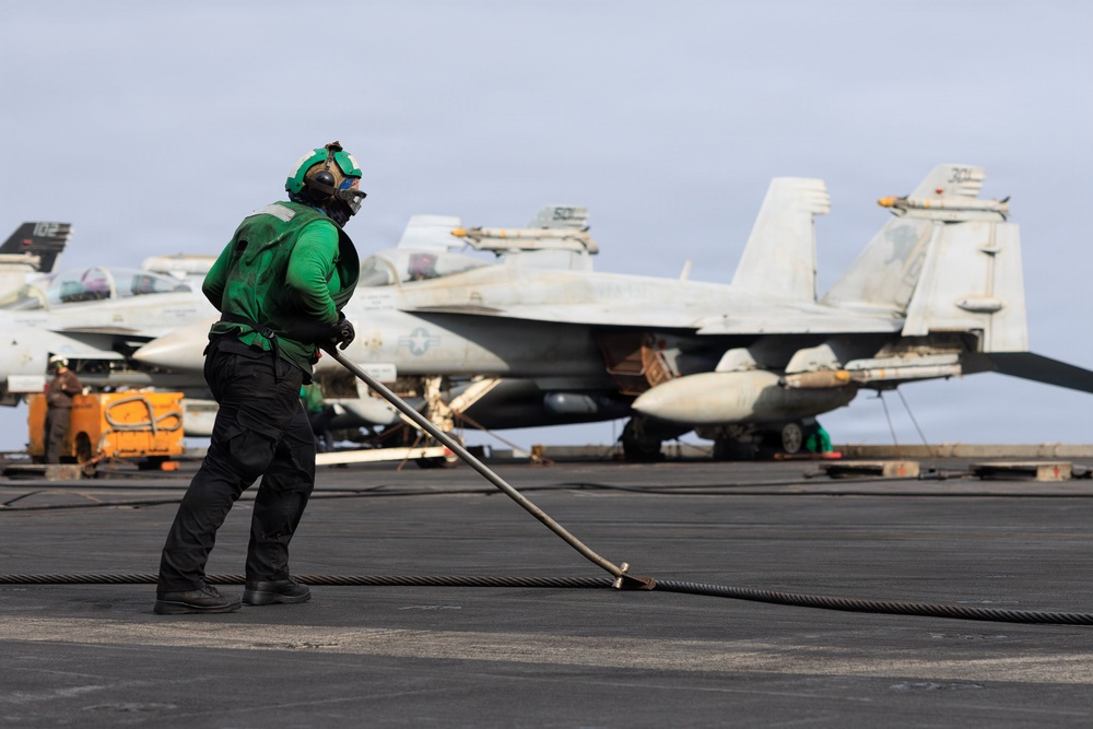 A U.S. Navy sailor in green jersey and cranial helmet works on the flight deck of an aircraft carrier, handling what appears to be an arresting wire or deck equipment with a long-handled tool. Multiple F/A-18 Hornet or Super Hornet jet aircraft are parked in the background, armed with missiles. The scene depicts routine flight deck operations aboard a U.S. Navy carrier.