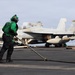 A U.S. Navy sailor in green jersey and cranial helmet works on the flight deck of an aircraft carrier, handling what appears to be an arresting wire or deck equipment with a long-handled tool. Multiple F/A-18 Hornet or Super Hornet jet aircraft are parked in the background, armed with missiles. The scene depicts routine flight deck operations aboard a U.S. Navy carrier.