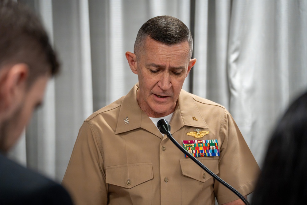 A senior U.S. Navy or Marine Corps officer in service khaki uniform is speaking at a microphone, appearing to read from notes or address an audience. The officer displays multiple service ribbons, a gold naval aviator or surface warfare badge, and collar insignia indicating senior rank. The setting appears to be an indoor formal or official event with a draped curtain backdrop.