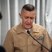 A senior U.S. Navy or Marine Corps officer in service khaki uniform is speaking at a microphone, appearing to read from notes or address an audience. The officer displays multiple service ribbons, a gold naval aviator or surface warfare badge, and collar insignia indicating senior rank. The setting appears to be an indoor formal or official event with a draped curtain backdrop.