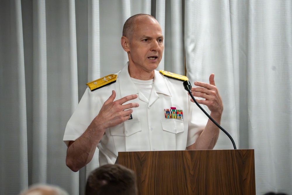 A senior military officer in white dress uniform is speaking at a wooden podium with a microphone. He is gesturing with both hands, suggesting an active and engaged speech or presentation. The setting appears to be an indoor formal or official event.