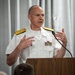 A senior military officer in white dress uniform is speaking at a wooden podium with a microphone. He is gesturing with both hands, suggesting an active and engaged speech or presentation. The setting appears to be an indoor formal or official event.