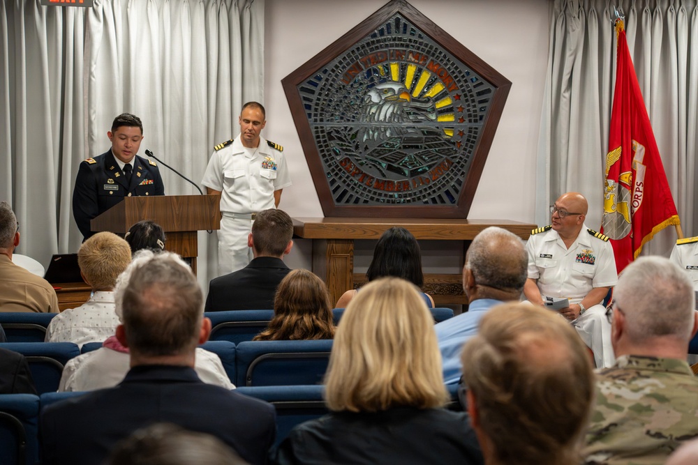 A formal military ceremony is taking place in what appears to be a chapel or ceremonial hall, likely at a U.S. military installation. An Army soldier in dress uniform is speaking at a podium while a Navy officer in dress whites stands nearby, with a seated audience of military personnel and civilians. A U.S. Marine Corps flag is visible in the background alongside a decorative stained-glass pentagon-shaped emblem.