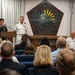 A formal military ceremony is taking place in what appears to be a chapel or ceremonial hall, likely at a U.S. military installation. An Army soldier in dress uniform is speaking at a podium while a Navy officer in dress whites stands nearby, with a seated audience of military personnel and civilians. A U.S. Marine Corps flag is visible in the background alongside a decorative stained-glass pentagon-shaped emblem.