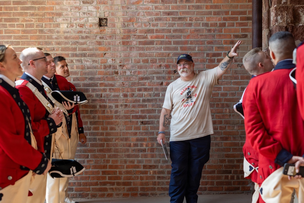 A man wearing a 'History Buff' t-shirt and baseball cap appears to be giving a tour or presentation to a group of individuals dressed in 18th-century British Redcoat military reenactment uniforms. The setting is against an exposed brick wall, suggesting a historic building or fort. The guide is gesturing with his raised hand, likely pointing out or explaining a historical feature.