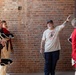A man wearing a 'History Buff' t-shirt and baseball cap appears to be giving a tour or presentation to a group of individuals dressed in 18th-century British Redcoat military reenactment uniforms. The setting is against an exposed brick wall, suggesting a historic building or fort. The guide is gesturing with his raised hand, likely pointing out or explaining a historical feature.