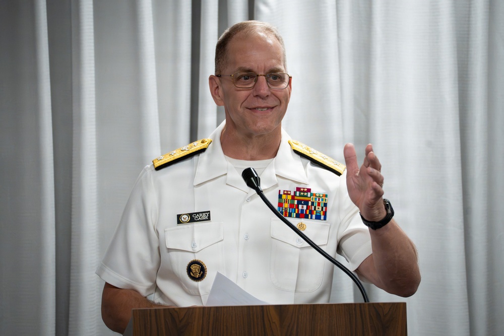 A U.S. Navy officer in white dress uniform is speaking at a wooden podium, gesturing with one hand. His name tag reads 'CAREY CASH' and he wears two-star rank insignia on his shoulders along with numerous service ribbons. He appears to be delivering a speech or presentation in front of a light gray curtain backdrop.