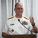 A U.S. Navy officer in white dress uniform is speaking at a wooden podium, gesturing with one hand. His name tag reads 'CAREY CASH' and he wears two-star rank insignia on his shoulders along with numerous service ribbons. He appears to be delivering a speech or presentation in front of a light gray curtain backdrop.