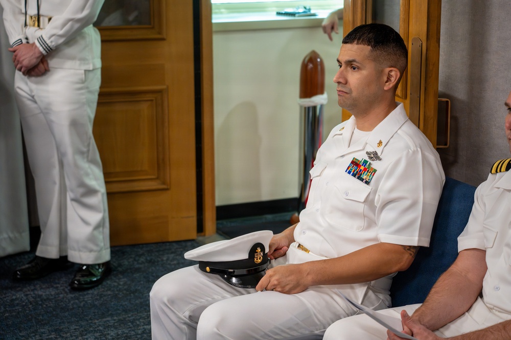 A U.S. Navy enlisted sailor in Service Dress White uniform is seated and holding his combination cover (white cap), appearing to attend a formal ceremony or official event. Multiple rows of service ribbons and a warfare device badge are visible on his chest. Other naval personnel in white uniforms are partially visible in the background and foreground.