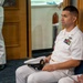 A U.S. Navy enlisted sailor in Service Dress White uniform is seated and holding his combination cover (white cap), appearing to attend a formal ceremony or official event. Multiple rows of service ribbons and a warfare device badge are visible on his chest. Other naval personnel in white uniforms are partially visible in the background and foreground.