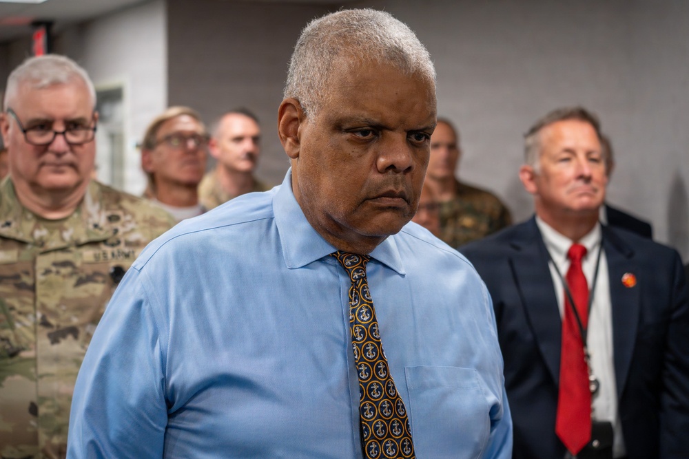 A group of individuals are gathered in what appears to be an indoor institutional setting. The central figure is a heavyset man in a light blue dress shirt and anchor-patterned tie with a serious expression. Behind him stand several people including individuals in U.S. Army camouflage uniforms and a man in a dark suit with a red tie.