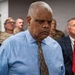 A group of individuals are gathered in what appears to be an indoor institutional setting. The central figure is a heavyset man in a light blue dress shirt and anchor-patterned tie with a serious expression. Behind him stand several people including individuals in U.S. Army camouflage uniforms and a man in a dark suit with a red tie.