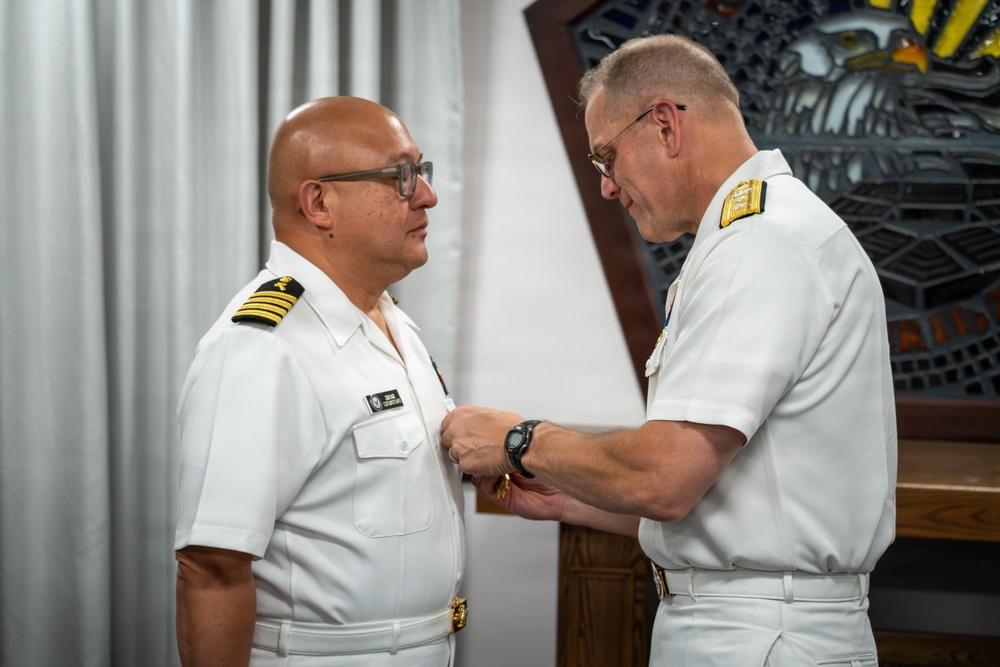 Two naval officers in white dress uniforms are engaged in a military decoration or pinning ceremony. The officer on the right is attaching or adjusting a medal or insignia on the chest of the officer on the left. Both wear gold shoulder boards indicating senior officer ranks.