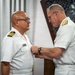 Two naval officers in white dress uniforms are engaged in a military decoration or pinning ceremony. The officer on the right is attaching or adjusting a medal or insignia on the chest of the officer on the left. Both wear gold shoulder boards indicating senior officer ranks.