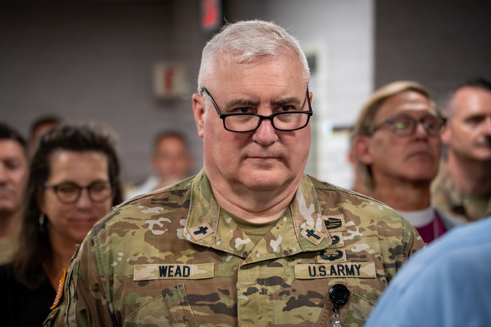 A U.S. Army soldier named Wead is prominently featured in the foreground, wearing OCP (Operational Camouflage Pattern) uniform with chaplain insignia (cross symbols) visible on the collar. He is wearing black-framed glasses and has a serious expression. Several civilians and at least one other religious figure are visible in the blurred background.
