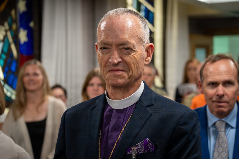 A clergy member wearing a purple clerical shirt with a white collar, dark blazer, and a gold pectoral cross stands in what appears to be a church interior. He has a serious expression and is wearing a purple pocket square. Several people are visible in the blurred background, along with stained glass windows.