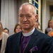 A clergy member wearing a purple clerical shirt with a white collar, dark blazer, and a gold pectoral cross stands in what appears to be a church interior. He has a serious expression and is wearing a purple pocket square. Several people are visible in the blurred background, along with stained glass windows.