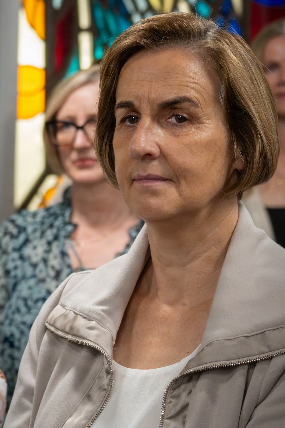 A middle-aged woman with short brown hair is in the foreground, wearing a light beige zip-up jacket over a white top. She appears to be attending a formal or ceremonial event inside a building with stained glass windows. Several other individuals are visible in the blurred background.