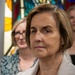 A middle-aged woman with short brown hair is in the foreground, wearing a light beige zip-up jacket over a white top. She appears to be attending a formal or ceremonial event inside a building with stained glass windows. Several other individuals are visible in the blurred background.