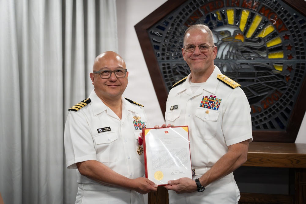 Two U.S. Navy officers in white dress uniforms are posing together while jointly holding a framed certificate or commendation document with a red border and gold seal. The officer on the left appears to be a Captain (O-6) based on visible rank insignia, and is wearing a medal on his chest, suggesting this is an awards or retirement ceremony. They are standing in front of a decorative Pentagon-shaped emblem with an eagle motif in the background.