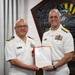 Two U.S. Navy officers in white dress uniforms are posing together while jointly holding a framed certificate or commendation document with a red border and gold seal. The officer on the left appears to be a Captain (O-6) based on visible rank insignia, and is wearing a medal on his chest, suggesting this is an awards or retirement ceremony. They are standing in front of a decorative Pentagon-shaped emblem with an eagle motif in the background.