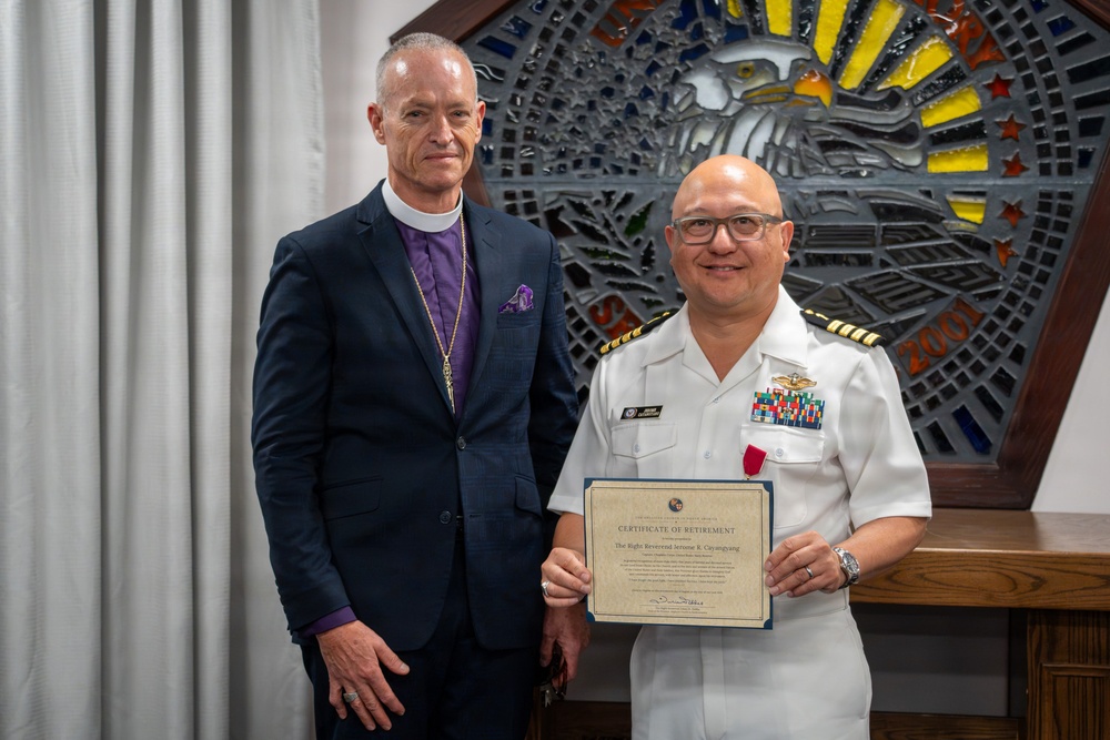 Two individuals pose together for a formal photograph, with one holding a Certificate of Retirement. The person on the right is wearing a white U.S. Navy officer's uniform with captain's insignia (four gold stripes), multiple service ribbons, and a medal. The person on the left is dressed in clerical attire with a purple shirt, pectoral cross, and episcopal collar, suggesting a bishop or senior clergy member.