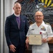Two individuals pose together for a formal photograph, with one holding a Certificate of Retirement. The person on the right is wearing a white U.S. Navy officer's uniform with captain's insignia (four gold stripes), multiple service ribbons, and a medal. The person on the left is dressed in clerical attire with a purple shirt, pectoral cross, and episcopal collar, suggesting a bishop or senior clergy member.