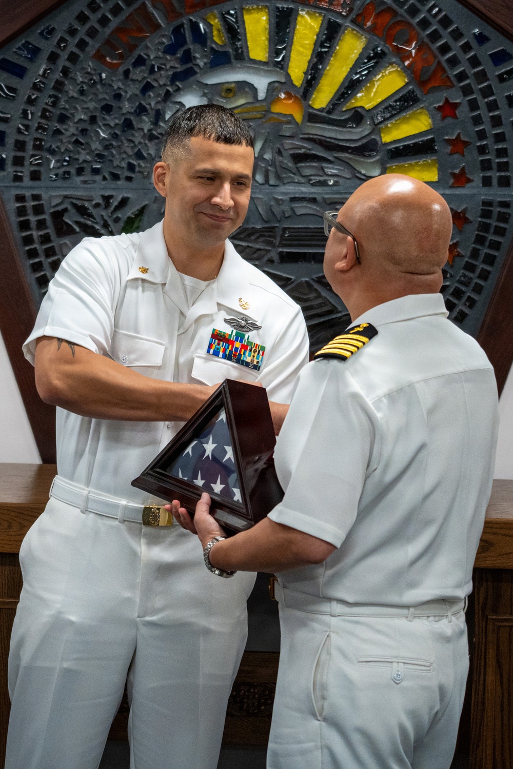 Two U.S. Navy personnel in white dress uniforms are exchanging a folded American flag in a triangular display case during what appears to be a military ceremony. The individual facing the camera wears multiple service ribbons, a warfare qualification badge, and collar insignia, while the other individual displays gold sleeve stripes indicating officer rank. The setting features a decorative stained-glass or mosaic eagle emblem in the background.