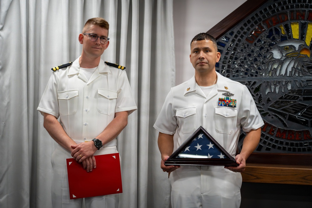 Two U.S. Navy personnel in white service uniforms are posed formally, likely during a military ceremony or retirement event. The individual on the right holds a triangularly folded American flag in a display case, while the individual on the left holds a red folder or certificate. A decorative military emblem or plaque is visible on the wall behind them.