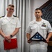 Two U.S. Navy personnel in white service uniforms are posed formally, likely during a military ceremony or retirement event. The individual on the right holds a triangularly folded American flag in a display case, while the individual on the left holds a red folder or certificate. A decorative military emblem or plaque is visible on the wall behind them.