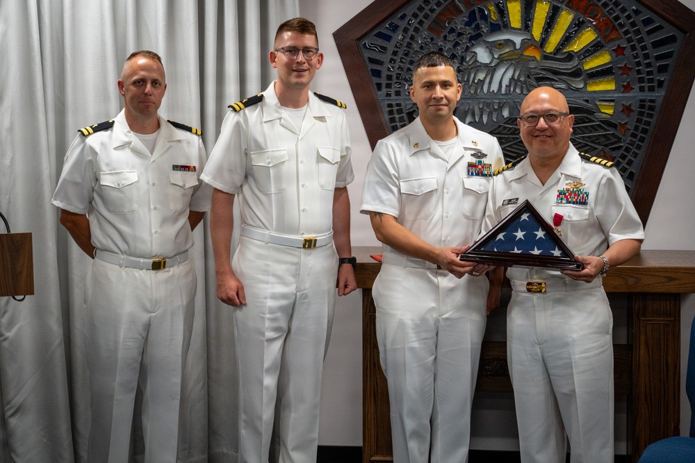 Four U.S. Navy personnel in white service dress uniforms are posed together during what appears to be a formal military ceremony. Two of the individuals are jointly holding a folded American flag in a triangular display case, suggesting a retirement or award ceremony. A decorative military insignia or seal is visible on the wall behind them.
