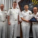 Four U.S. Navy personnel in white service dress uniforms are posed together during what appears to be a formal military ceremony. Two of the individuals are jointly holding a folded American flag in a triangular display case, suggesting a retirement or award ceremony. A decorative military insignia or seal is visible on the wall behind them.