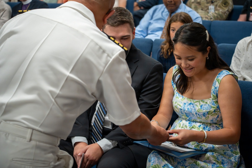A naval officer in white dress uniform is leaning forward to interact with a smiling young woman in a floral dress who is seated in a blue auditorium chair. A young man in a dark suit and striped tie is seated beside her. The setting appears to be a formal military ceremony or presentation event.