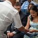 A naval officer in white dress uniform is leaning forward to interact with a smiling young woman in a floral dress who is seated in a blue auditorium chair. A young man in a dark suit and striped tie is seated beside her. The setting appears to be a formal military ceremony or presentation event.