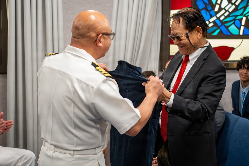 A military or naval officer in a white uniform with gold shoulder insignia is presenting a dark garment to a civilian man wearing a black suit and red tie. The civilian appears pleased, smiling during the exchange. The setting appears to be an indoor ceremony or formal event, with stained glass visible in the background.