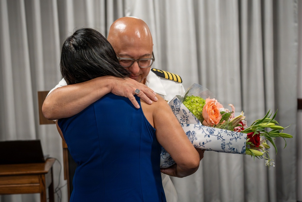 A man in a white military or naval dress uniform with gold rank insignia on the shoulder is embracing a woman in a blue dress. The woman is holding a bouquet of flowers wrapped in decorative blue and white floral paper. The moment appears to be an emotional or celebratory occasion, possibly a ceremony or formal event.