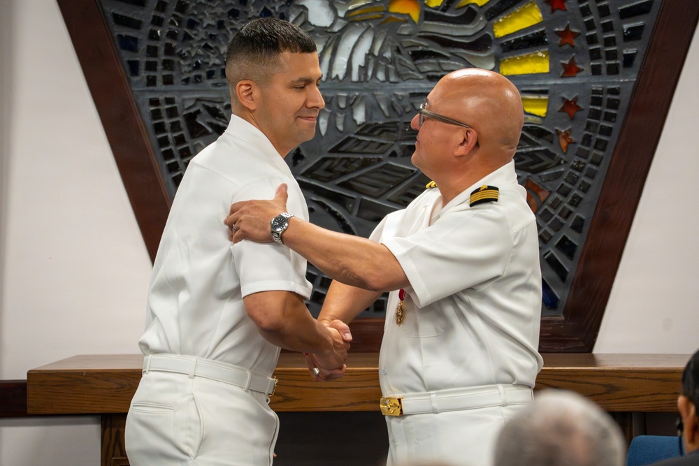 Two naval officers in white dress uniforms are engaged in what appears to be a formal military ceremony, likely a promotion or award pinning. The officer on the right, who has gold shoulder boards indicating senior rank, is placing or adjusting something on the shoulder or collar of the officer on the left. The setting appears to be an indoor military chapel or ceremonial hall with a decorative mosaic or stained glass backdrop.